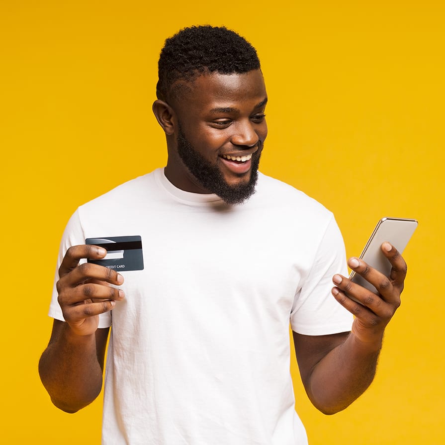 Cheerful black man using credit card and smartphone for purchasing online happy guy looking at phone and holding debit card in other hand