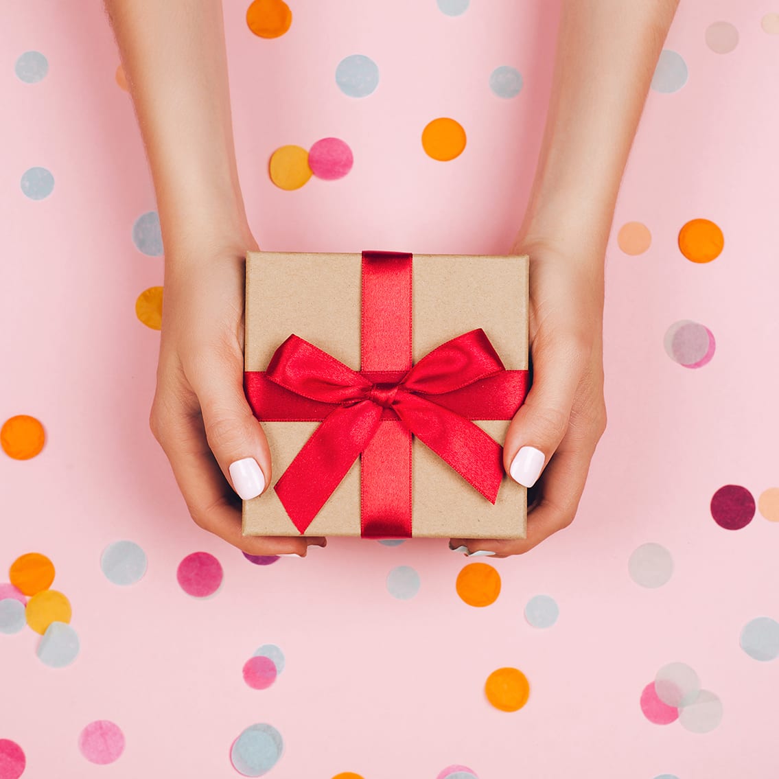 Woman hands holding present box with red bow on pastel pink background with multicolored confetti. Flat lay style.