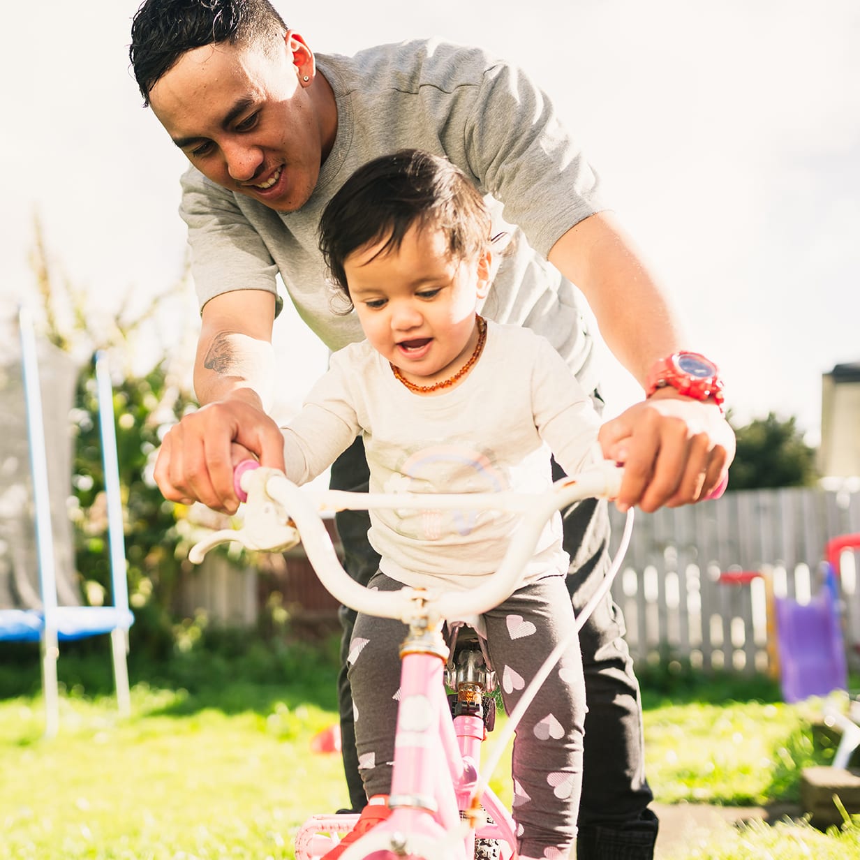 Families From New Zealand. Maori father helping her daughter to ride bicycle in backyard.