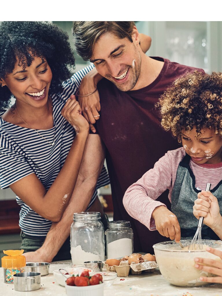 A mom, dad, and child are in the kitchen. The mom and dad stand smiling behind the child who is mixing dough in a mixing bowl
