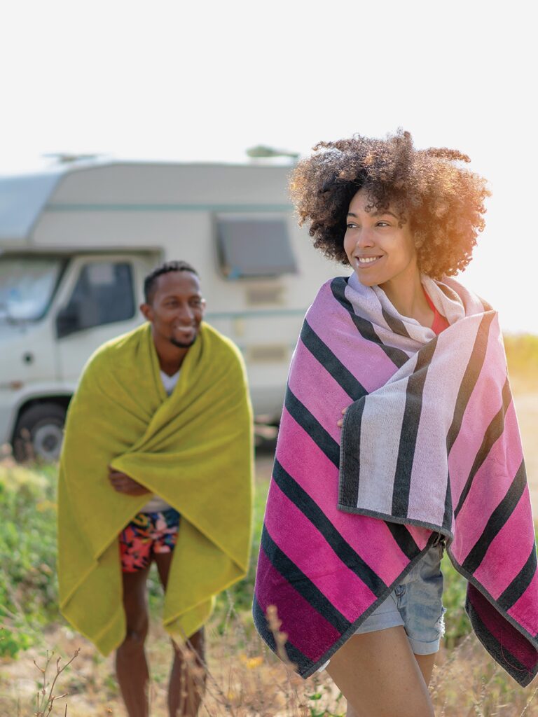 Photo of a woman and a man outside. They are wearing summer clothes and have beach towels wrapped around their shoulders. There is an RV in the background.
