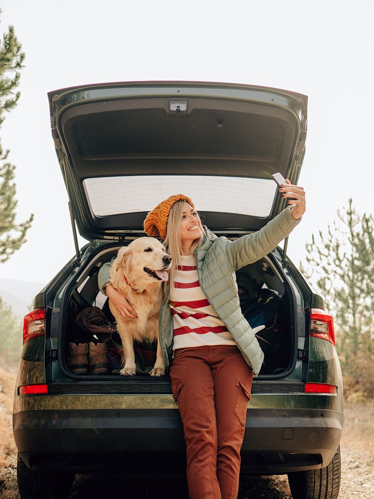 A woman and her dog leaning on the open tailgate of her car. She is taking a photo of herself and her dog.