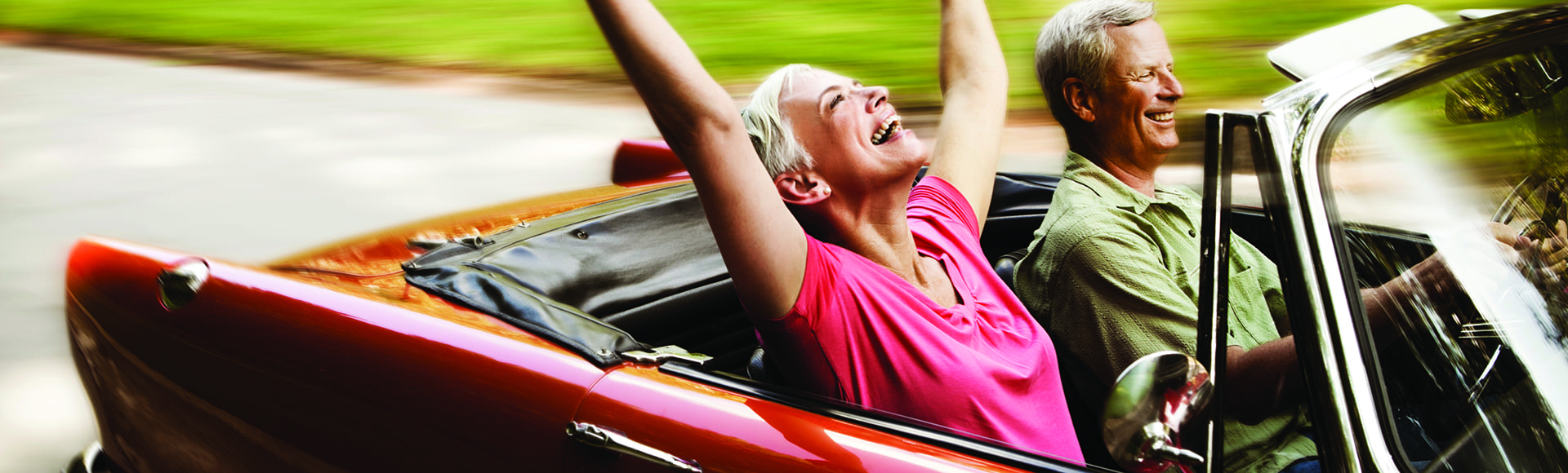 Senior woman throws her arms in the air while her husband drives her down a road in their convertible. Horizontal shot.