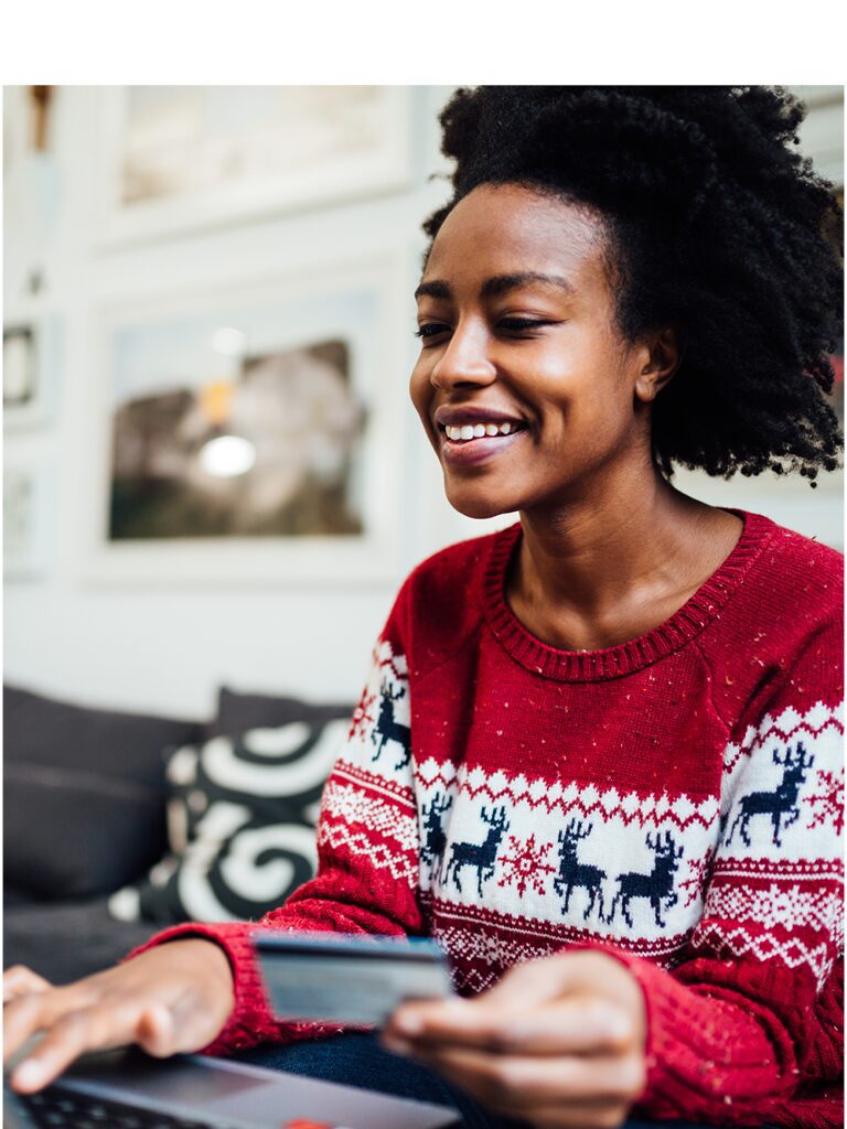 Image of a smiling woman in a red festive sweater - she is holding her credit card and typing on a computer keyboard