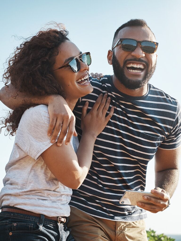 zeal-Credit-Card-Campaign-SUMMER-photo-768x1024 A man and a woman are smiling and laughing. He has his arm wrapped around her shoulder. They are outside on a sunny day.