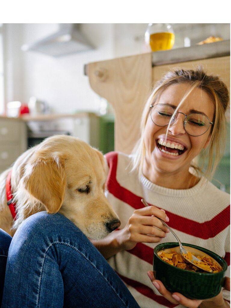 A woman is smiling and laughing as her golden retriever stares eagerly at the bowl of food in the woman's hands