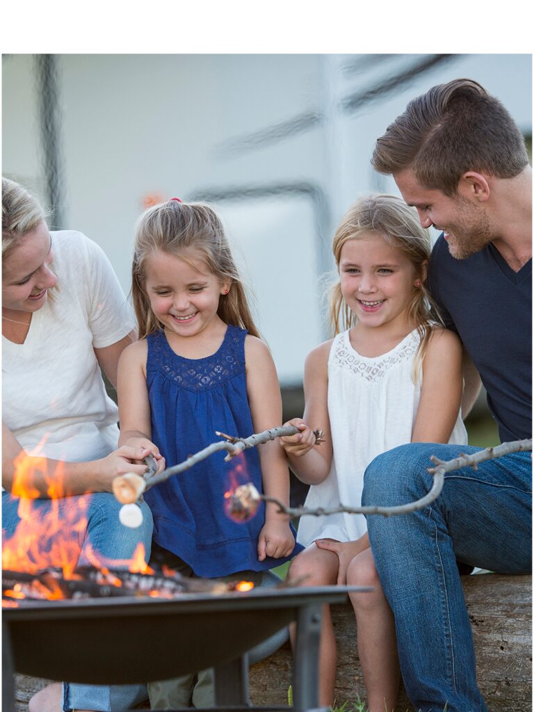 Two young girls are between their mother on the left and their father on the right. The family is seated in front of a fire pit and they are happily toasting marshmallows.